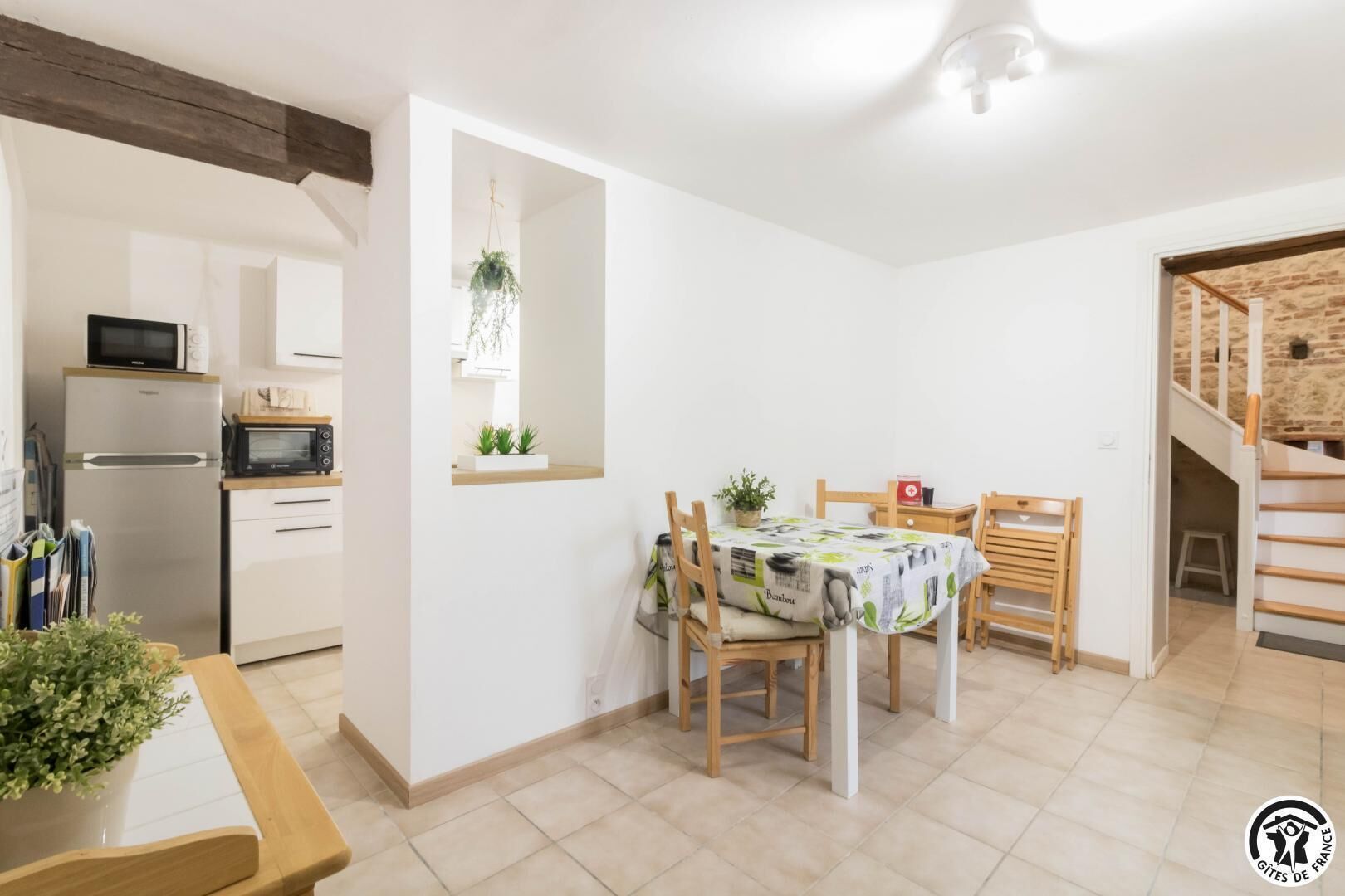 A compact kitchen with a refrigerator, microwave, and wooden cabinets. A dining area with a table and chairs is adjacent, featuring a tablecloth and potted plants.