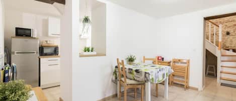 A compact kitchen with a refrigerator, microwave, and wooden cabinets. A dining area with a table and chairs is adjacent, featuring a tablecloth and potted plants.