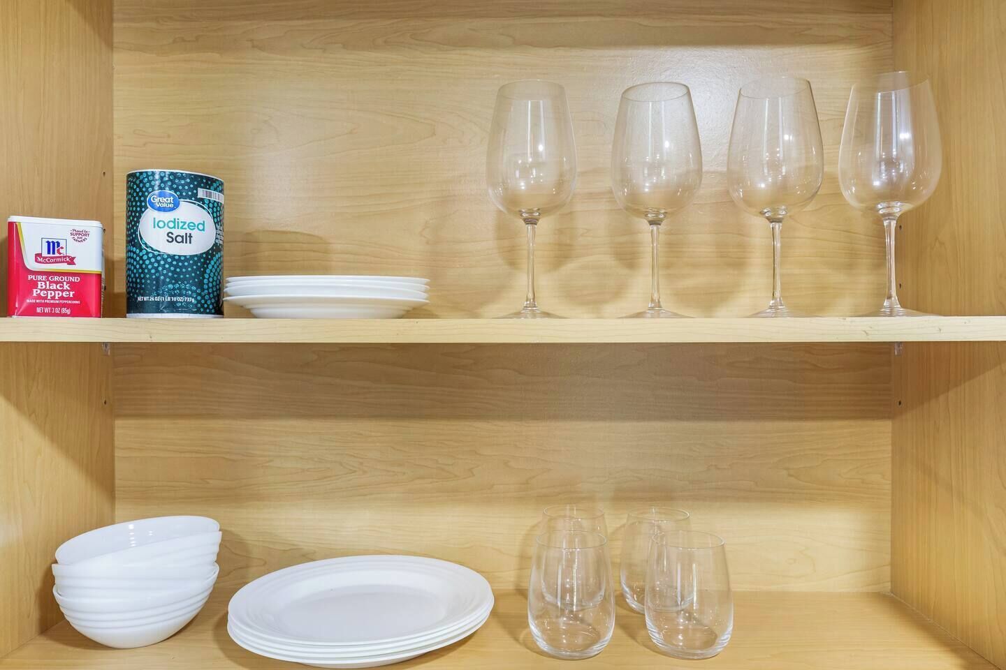 A wooden shelf with white plates, clear wine glasses, a can of iodized salt, and a box of McCormick black pepper.