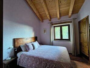 A bedroom with a wooden ceiling, a bed with patterned bedding, a bedside table with a lamp, a window with curtains, and a wardrobe.
