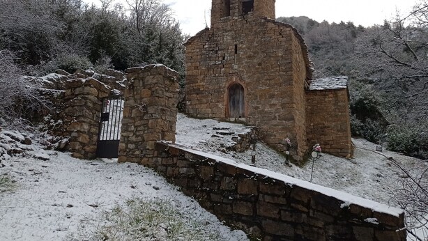 Un edifici antic de pedra amb una torre campanar, envoltat d'arbres coberts de neu i una paret de pedra.