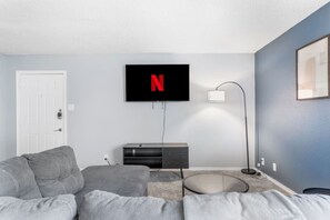 Ein modernes Wohnzimmer mit grauem Sofa, einem an der Wand befestigten Fernseher, einem Beistelltisch und einer Lampe.