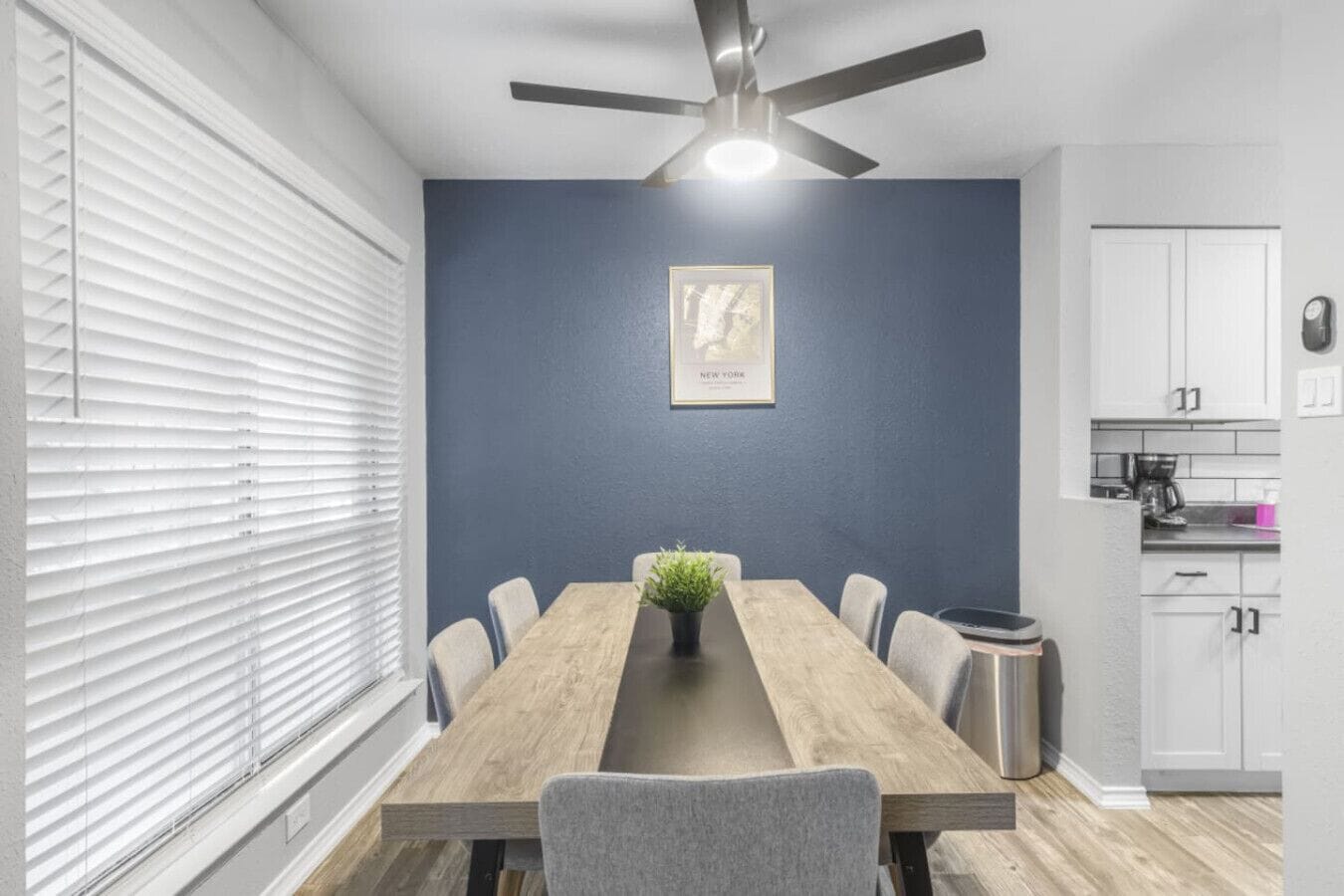 A dining area with a wooden table, grey chairs, a ceiling fan, and a kitchen area with cabinets and appliances.