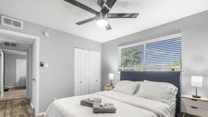 A bedroom with a bed, a ceiling fan, a window with blinds, and a nightstand with a lamp. - Arlington 3BR | AT&T, Globe Life, 6 Flags, Parking (Arlington)