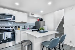A modern kitchen with white cabinetry, stainless steel appliances, a central island, and a staircase leading to another level. - 5 Min to Love Field | Gated+Guard | 2BR | Parking (Dallas)