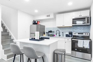 A modern kitchen with white cabinetry, a stainless steel range, a microwave, and a built-in oven. There are two grey chairs and a small trash bin. - 5 Min to Love Field | Gated+Guard | 2BR | Parking (Dallas)