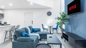 En moderne stue med en hjørnesofa, et fjernsyn monteret på væggen, et spiseområde med stole og et lille køkken med en mikrobølgeovn.