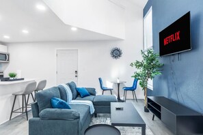 A modern living room with a sectional sofa, a TV mounted on the wall, a dining area with chairs, and a small kitchen area with a microwave. - 5 Min to Love Field | Gated+Guard | 2BR | Parking (Dallas)
