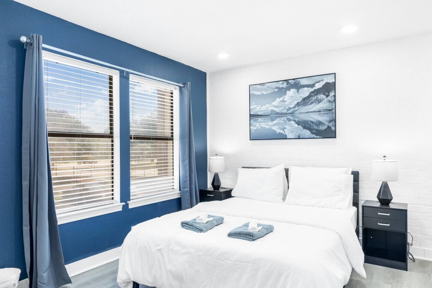 A bedroom with a bed, two bedside tables, a window with blinds, and a wall-mounted picture.