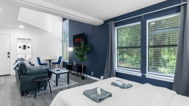 A modern hotel room with a bed, seating area, TV, and large windows. - 5 Min to Love Field | Gated+Guard | 2BR | Parking (Dallas)