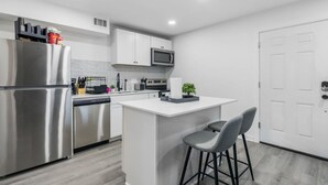 A modern kitchen with a stainless steel refrigerator, a microwave, and a built-in oven. There is a small island with a bar stool and a potted plant. - 5 Min to Love Field | Gated+Guard | 2BR | Parking (Dallas)