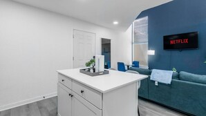 A modern living room with a white cabinet, a blue sofa, a TV displaying 'NETFLIX,' and a small plant on the cabinet. - 5 Min to Love Field | Gated+Guard | 2BR | Parking (Dallas)