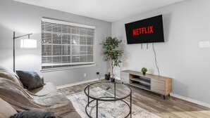 A living room with a sofa, a coffee table, a TV mounted on the wall, a window with blinds, and a plant. - Arlington 1BR | AT&T, Globe Life, 6 Flags, Parking (Arlington)