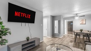 A modern living room with a mounted TV displaying 'NETFLIX,' a dining area with a table and chairs, and a hallway leading to another room. - Arlington 1BR | AT&T, Globe Life, 6 Flags, Parking (Arlington)