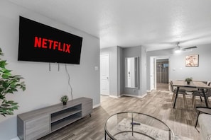 A modern living room with a mounted TV displaying 'NETFLIX,' a dining area with a table and chairs, and a hallway leading to another room. - Arlington 1BR | AT&T, Globe Life, 6 Flags, Parking (Arlington)