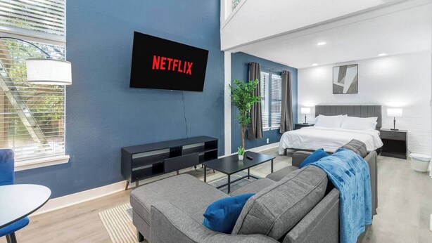 A modern hotel room with a large bed, a TV mounted on the wall, a seating area with a sofa and a small table, and a window with blinds. - 5 Min to Love Field | Gated+Guard | 2BR | Parking (Dallas)