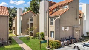 A residential building with a tiled roof, a green lawn, and a parking area with cars. - 5 Min to Love Field | Gated+Guard | 2BR | Parking (Dallas)