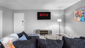 A modern living room with a flat-screen TV mounted on the wall, a sofa with cushions, a glass coffee table, and a painting on the wall. - Arlington 2BR | AT&T, Globe Life, 6 Flags, Parking (Arlington)