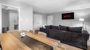 A modern living room with a wooden dining table, a dark grey sofa, a flat-screen TV mounted on the wall, and a ceiling fan. - Arlington 2BR | AT&T, Globe Life, 6 Flags, Parking (Arlington)