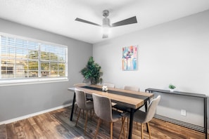 Un coin repas avec une table en bois, quatre chaises, un ventilateur de plafond, une plante en pot et un tableau accroché au mur.