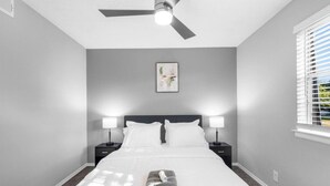 A bedroom with a bed, two bedside lamps, a ceiling fan, a window with blinds, and a framed picture on the wall. - Arlington 2BR | AT&T, Globe Life, 6 Flags, Parking (Arlington)