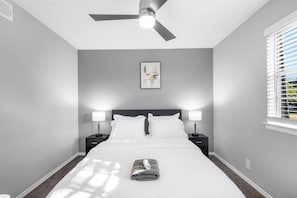 Une chambre à coucher comprenant un lit, deux lampes de chevet, un ventilateur de plafond, une fenêtre avec des stores et un tableau encadré accroché au mur.