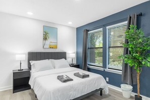 A modern bedroom with a bed, bedside tables, a nightstand, a window with blinds, a plant, and a painting on the wall. - 5 Min to Love Field | Gated+Guard | 2BR | Parking (Dallas)