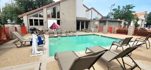 A swimming pool with lounge chairs, a lifeguard stand, and a building with large windows and a red roof. - 5 Min to Love Field | Gated+Guard | 2BR | Parking (Dallas)