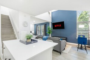A modern living room with a white central island, grey sofas, a blue accent chair, a TV mounted on a blue wall, and a staircase leading to another level. - 5 Min to Love Field | Gated+Guard | 2BR | Parking (Dallas)