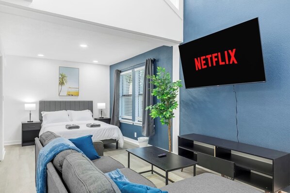 A modern hotel room with a bed, a sofa, a TV mounted on the wall, and a plant. - 5 Min to Love Field | Gated+Guard | 2BR | Parking (Dallas)