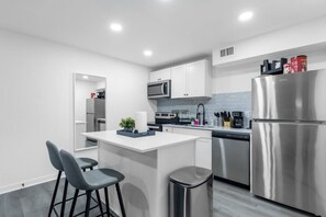Una cocina moderna con armarios blancos, electrodomésticos de acero inoxidable, una isla central con taburetes y un cubo de basura.