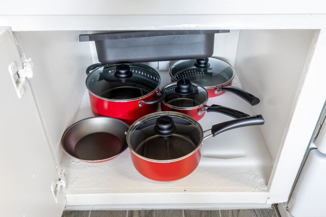 Ett köksskåp med fyra röda kastruller och stekpannor, ett svart lock och en vit bänkskiva.