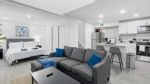 Una habitación de hotel moderna con cama, sofá, mesa de centro, kitchenette y un cuadro en la pared.