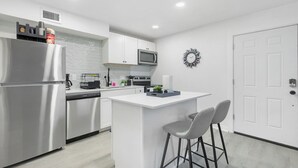 Una cocina moderna con refrigerador de acero inoxidable, microondas y lavavajillas, una encimera blanca y una pequeña isla con dos taburetes de bar.