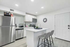 A modern kitchen with a stainless steel refrigerator, microwave, and dishwasher, a white countertop, and a small island with two bar stools. - 5 Min to Love Field | Gated+Guard | 2BR | Parking (Dallas)