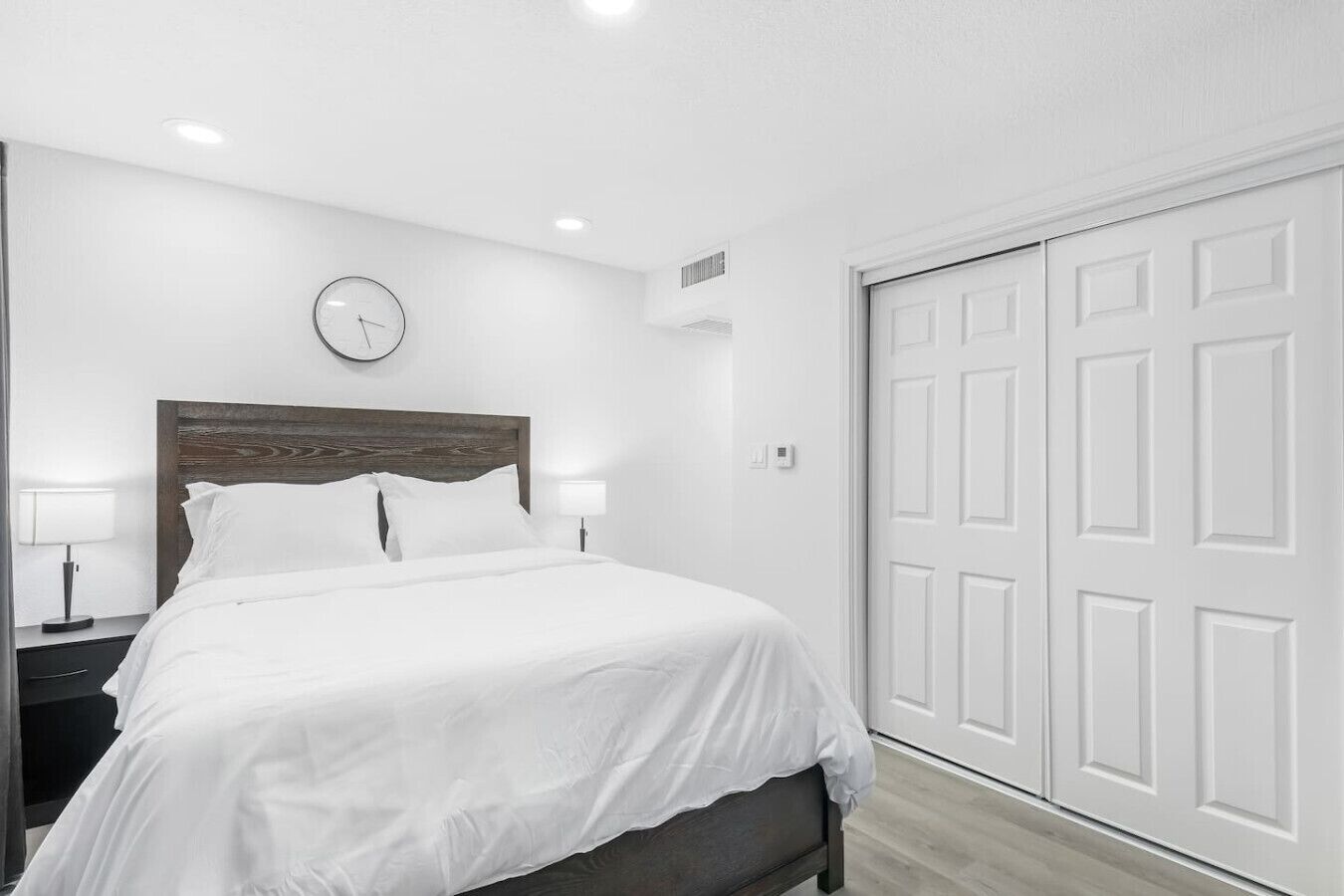 A hotel room with a bed, bedside lamps, a clock, and a white wardrobe.