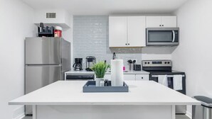 Une cuisine moderne avec des meubles blancs, un réfrigérateur en acier inoxydable, un four à micro-ondes, une machine à café et un îlot central agrémenté d’une plante et d’un plateau.