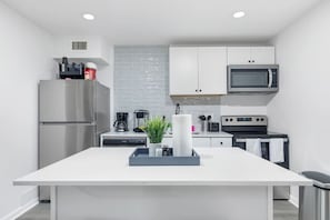 A modern kitchen with white cabinetry, a stainless steel refrigerator, a microwave, a coffee maker, and a central island with a plant and a tray.
