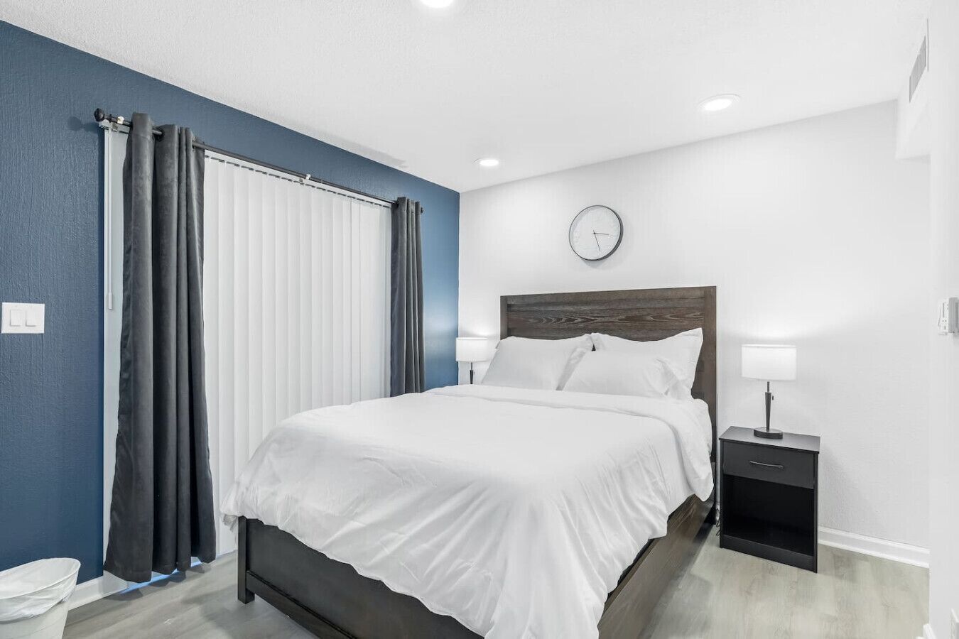 Une chambre à coucher moderne avec un lit, une table de chevet, une horloge murale et une fenêtre avec des rideaux.