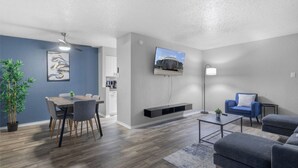 A modern living room with a dining area, a flat-screen TV mounted on the wall, a ceiling fan, and a potted plant.