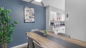 A dining area with a wooden table and chairs, a ceiling fan, a wall-mounted abstract painting, and a kitchen in the background.