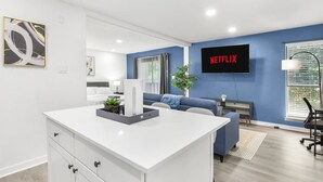 A modern living room with a white entertainment center, a blue sofa, a TV mounted on the wall, and a potted plant. - 5 Min to Love Field | Gated+Guard | 1BR | Parking (Dallas)