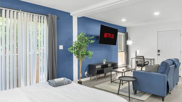 A modern hotel room with a bed, a seating area with a sofa and a small table, a TV mounted on the wall, and a potted plant. - 5 Min to Love Field | Gated+Guard | 1BR | Parking (Dallas)