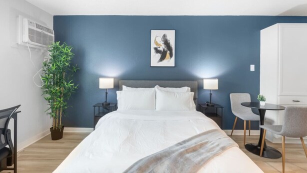A modern hotel room with a large bed, two bedside tables, a desk with a chair, and a potted plant. - Fort Worth Studio3 – 10 Min to Downtown, Gym, Pets (Haltom City)