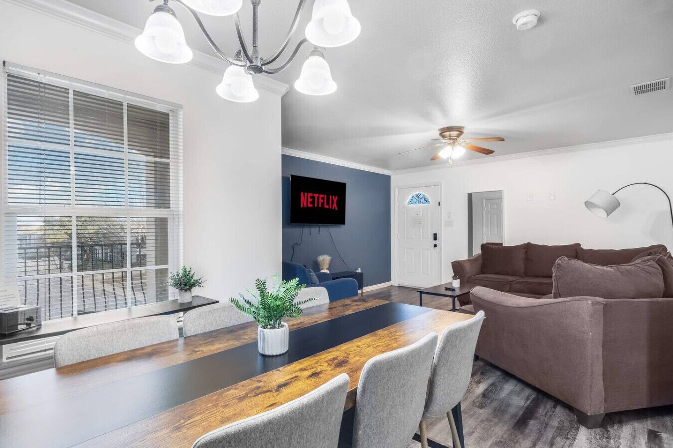 Een moderne woonkamer met een houten eettafel, grijze banken, een plafondventilator en een tv met daarop 'NETFLIX'.
