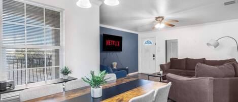 Een moderne woonkamer met een houten eettafel, grijze banken, een plafondventilator en een tv met daarop 'NETFLIX'.