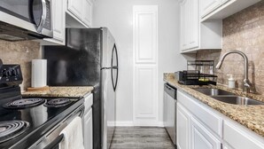 Una cocina equipada con estufa, microondas, refrigerador, encimera de granito, pileta de acero inoxidable y alacenas blancas.