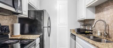A kitchen with a stove, microwave, refrigerator, granite countertop, stainless steel sink, and white cabinetry.