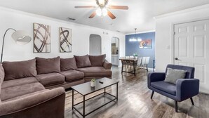 Una sala de estar con un sofá modular, una mesita de centro, un sillón azul y una zona de comedor con mesa y sillas.