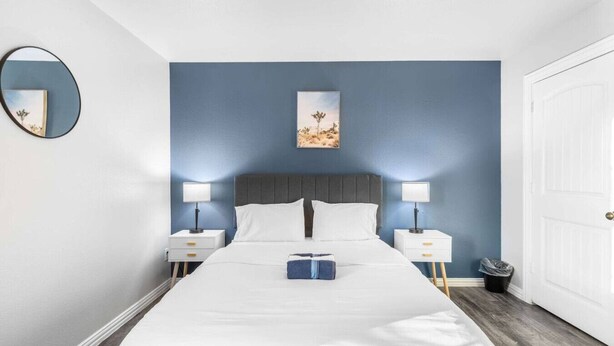 Un dormitorio con una cama, dos mesitas de noche, una mesita auxiliar, un cuadro en la pared y una puerta.
